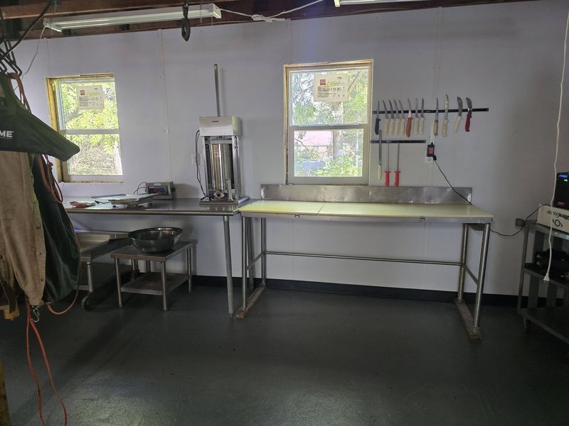 Processing room with stainless-steel tables, a sausage stuffer, a digital scale and a wall-mounted knife rack between two windows